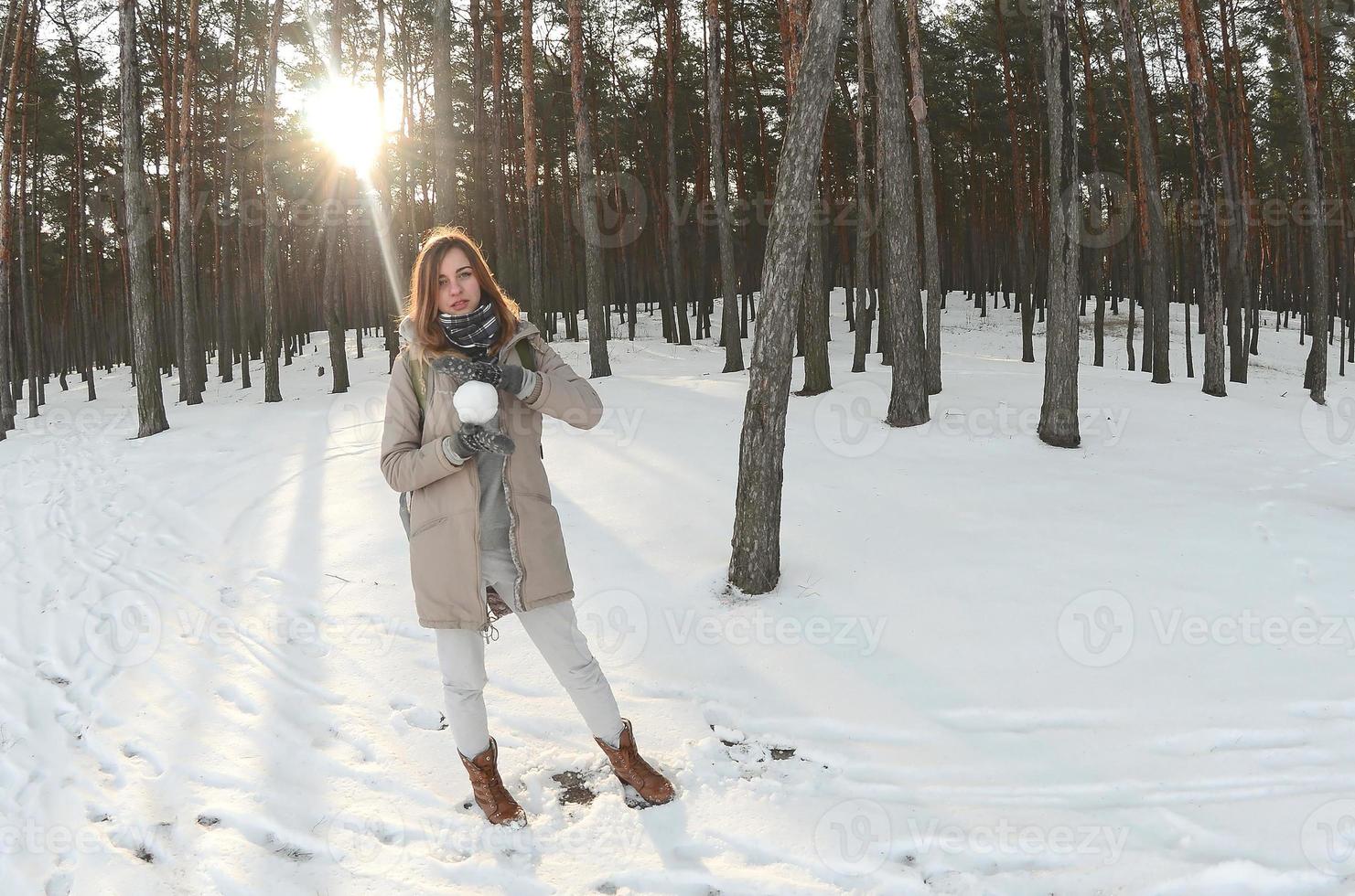 A young and joyful Caucasian girl in a brown coat holds a snowball in a snow-covered forest in winter. Fisheye Photo