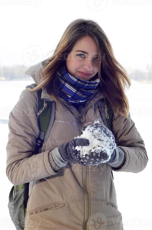 A young and joyful Caucasian girl in a brown coat holds a snowball in the background of a horizon line between the sky and a frozen lake in winter photo
