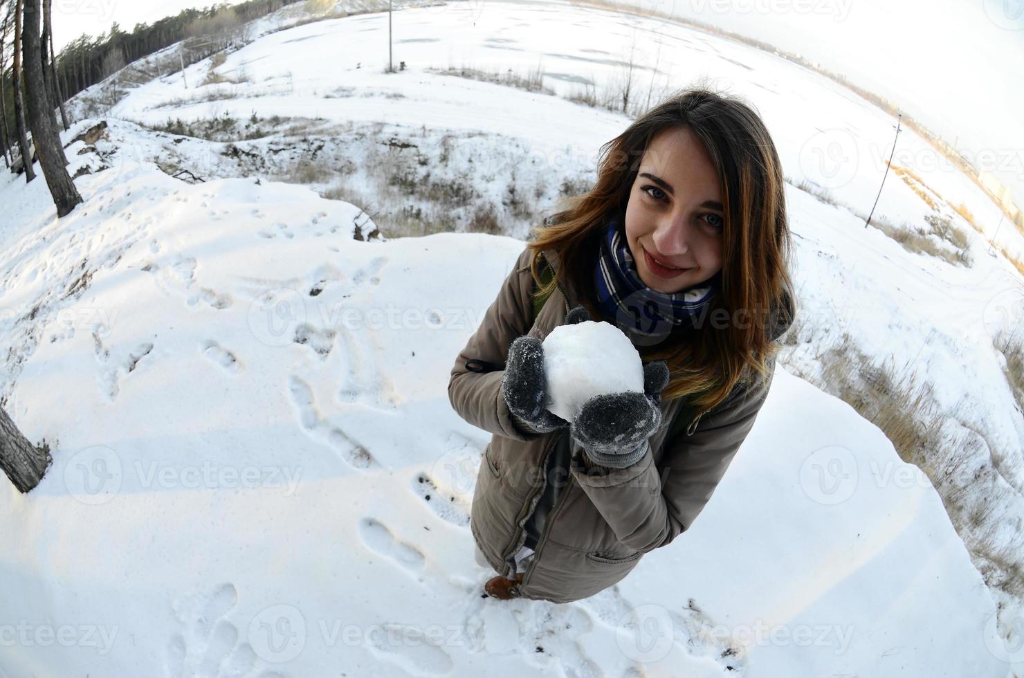 A young and joyful Caucasian girl in a brown coat holds a snowball in front of a horizon line between the sky and a frozen lake in winter. Fisheye Photo