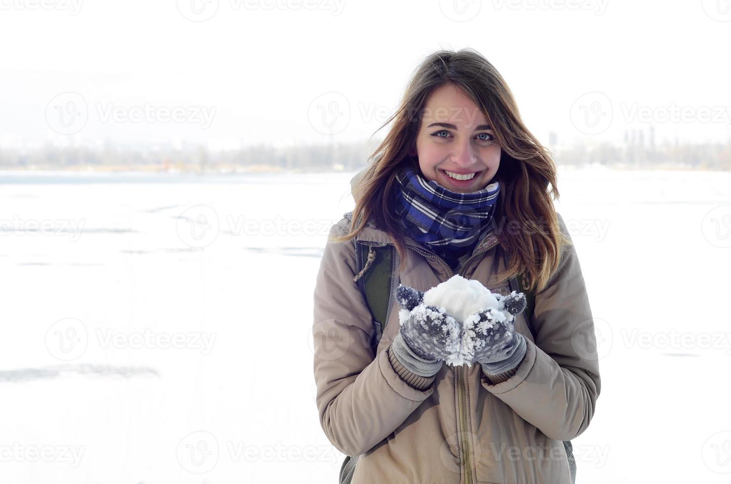 A young and joyful Caucasian girl in a brown coat holds a snowball in the background of a horizon line between the sky and a frozen lake in winter photo