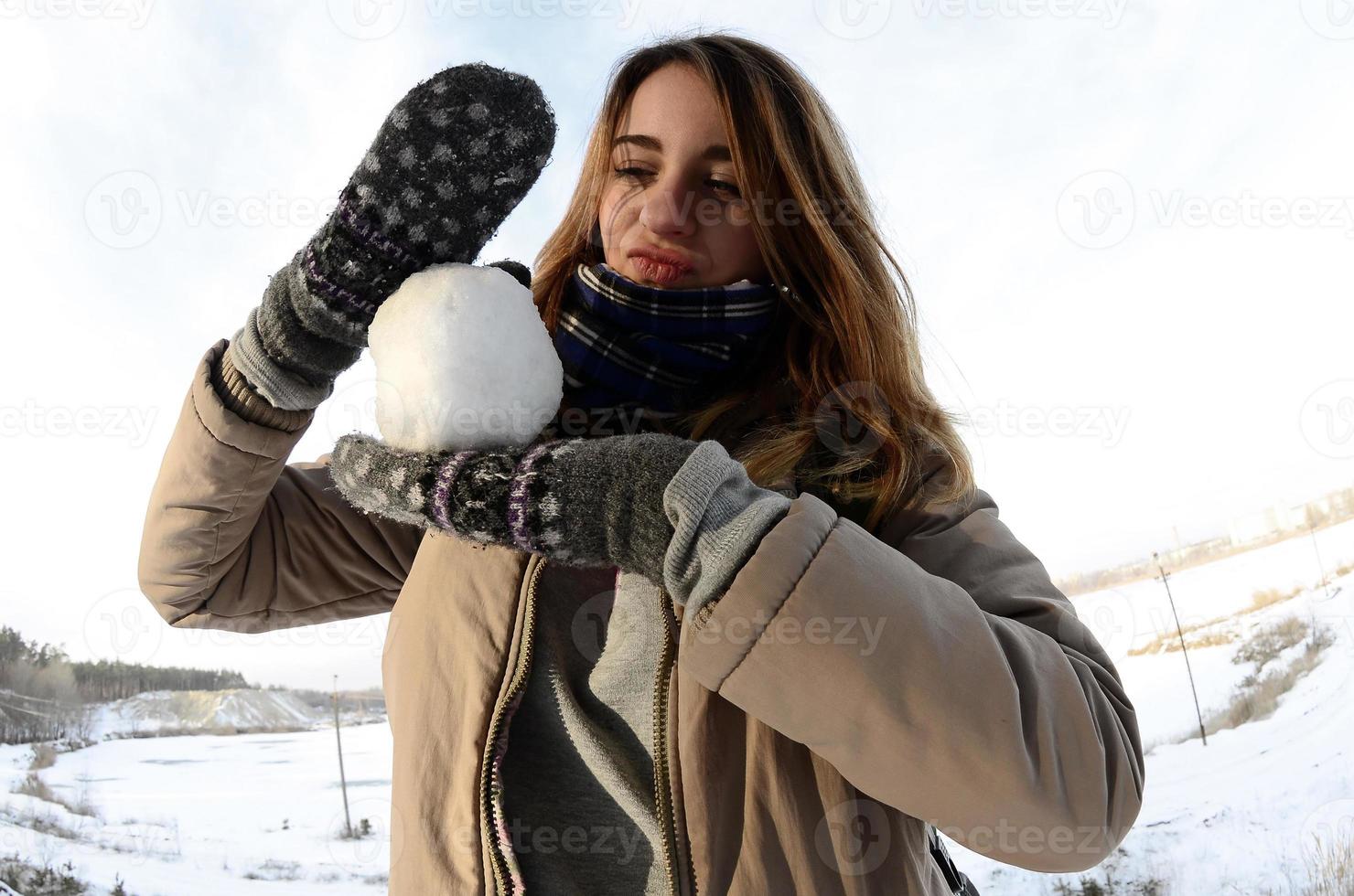 A young and joyful Caucasian girl in a brown coat holds a snowball in front of a horizon line between the sky and a frozen lake in winter. Fisheye Photo