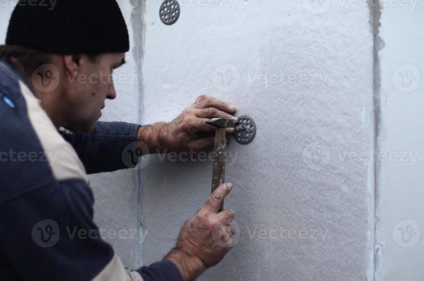 un trabajador anciano obstruye una espiga en un paraguas de plástico montado en una pared de espuma de poliestireno. el proceso de fijación de placas de poliestireno expandido. calentamiento de la fachada del edificio foto