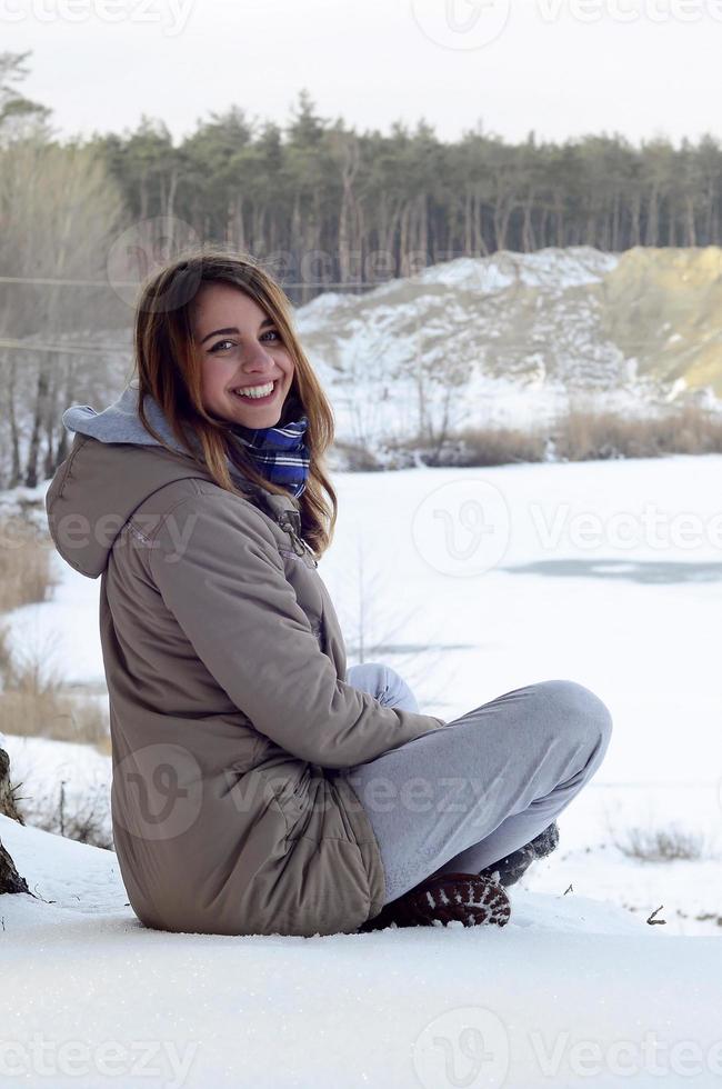 A young Caucasian girl in a brown coat is sitting near a cliff in the background of a horizon line between the sky and a frozen lake in winter time photo