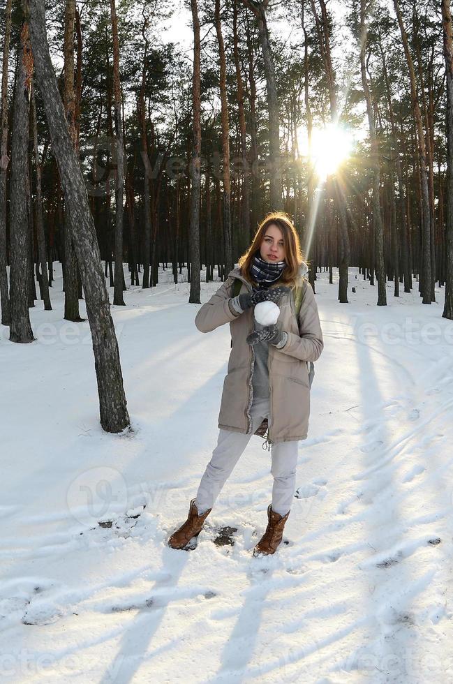 A young and joyful Caucasian girl in a brown coat holds a snowball in a snow-covered forest in winter. Fisheye Photo