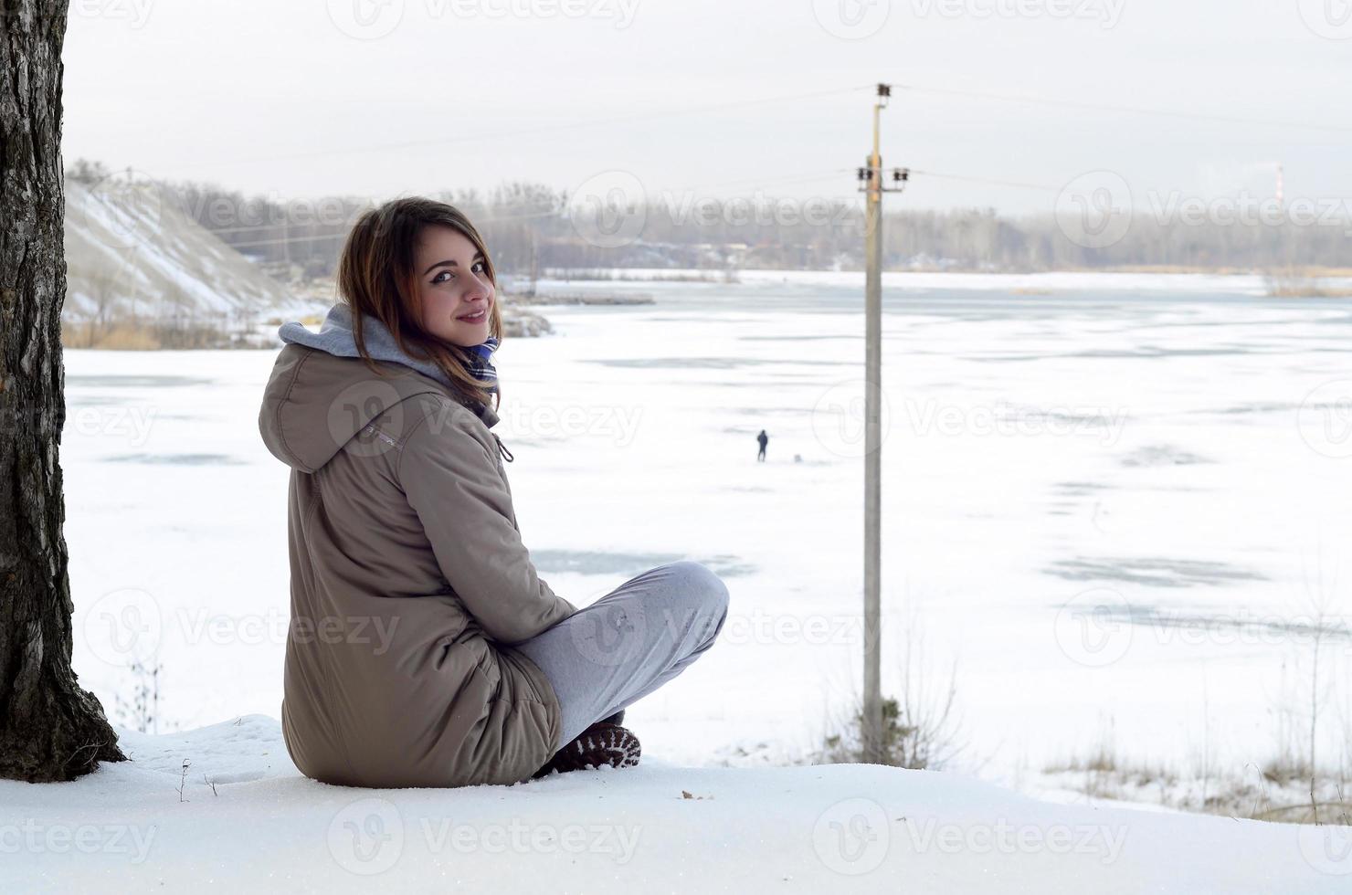 A young Caucasian girl in a brown coat is sitting near a cliff in the background of a horizon line between the sky and a frozen lake in winter time photo