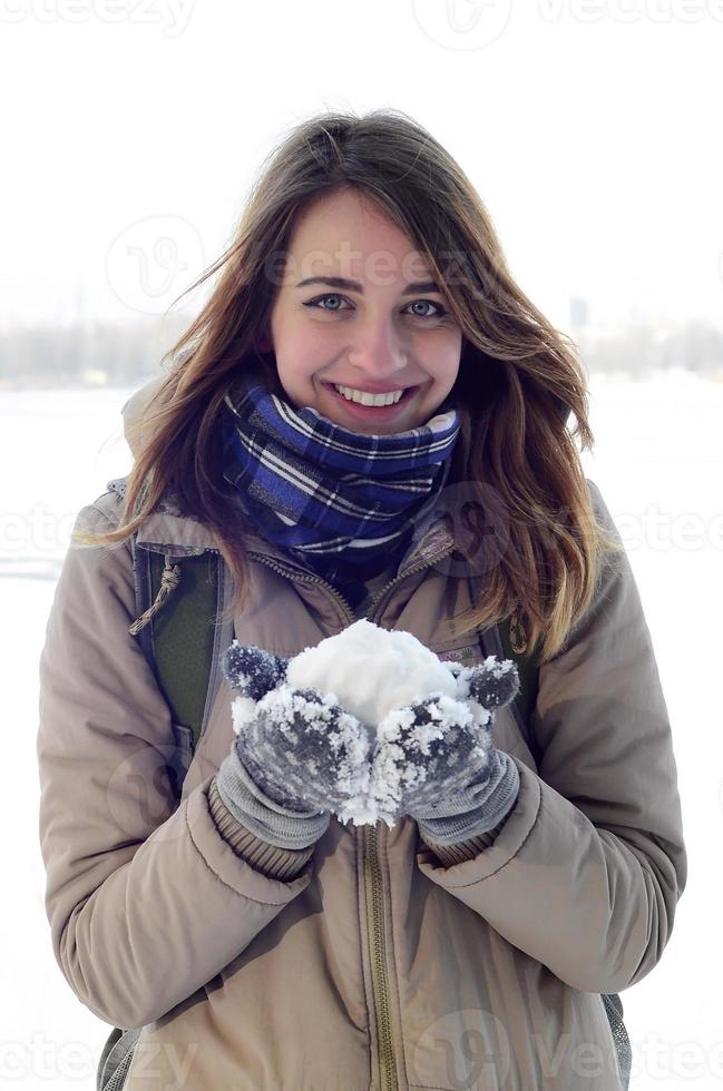 A young and joyful Caucasian girl in a brown coat holds a snowball in the background of a horizon line between the sky and a frozen lake in winter photo