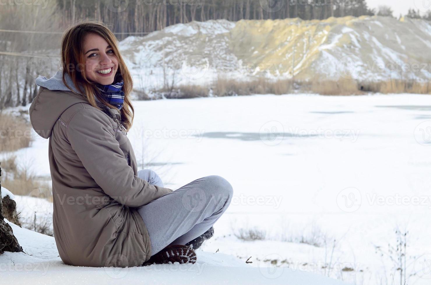 A young Caucasian girl in a brown coat is sitting near a cliff in the background of a horizon line between the sky and a frozen lake in winter time photo