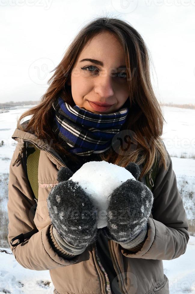 una joven y alegre chica caucásica con un abrigo marrón sostiene una bola de nieve frente a una línea de horizonte entre el cielo y un lago congelado en invierno. foto de ojo de pez