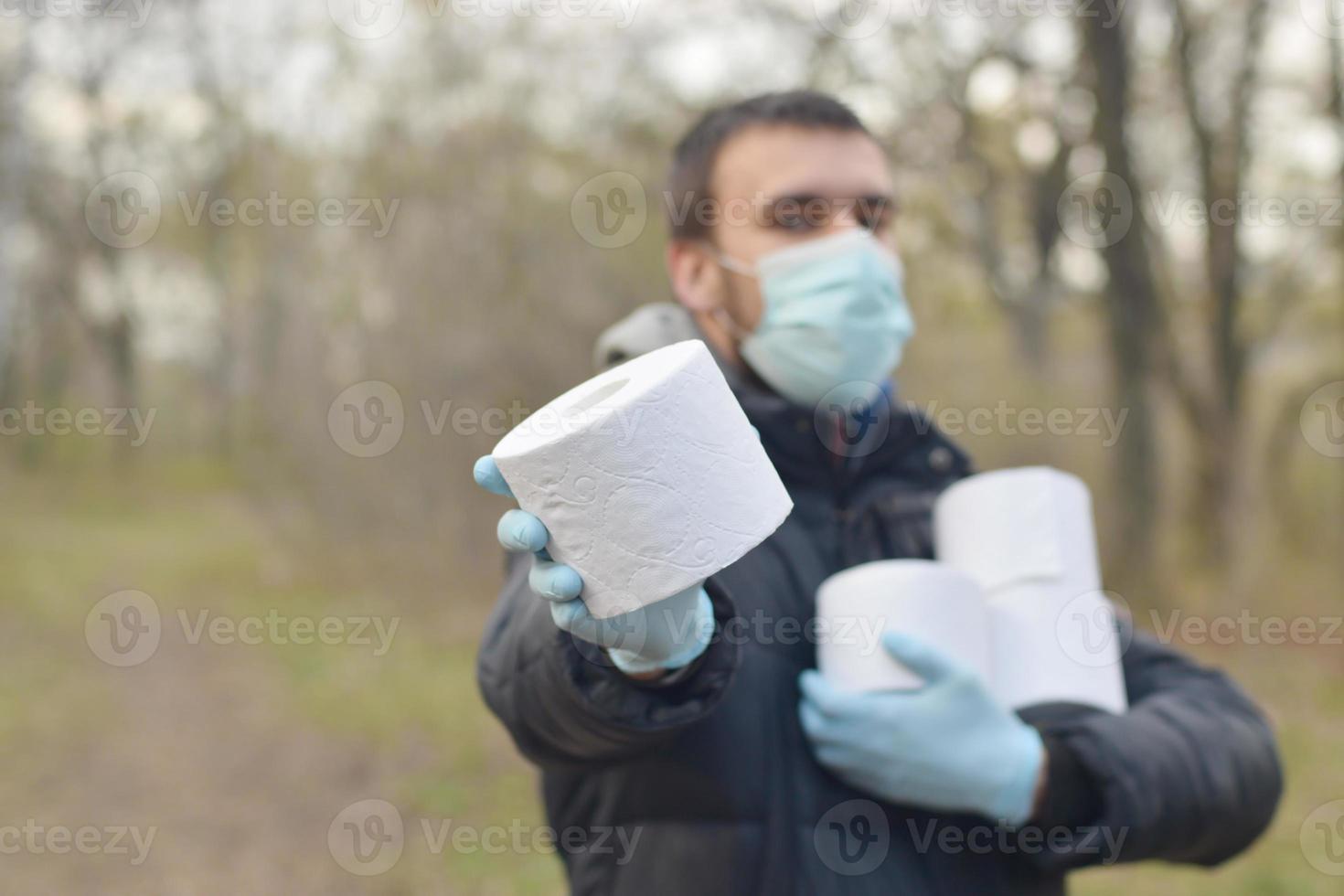 Covidiot concept. Young man in protective mask holds many rolls of toilet paper outdoors in spring wood photo