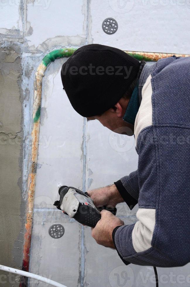 un trabajador anciano perfora un agujero en una pared de espuma de poliestireno para la posterior instalación de una espiga de refuerzo de plástico. creando agujeros en la pared con un taladro. calentamiento de la fachada del edificio foto