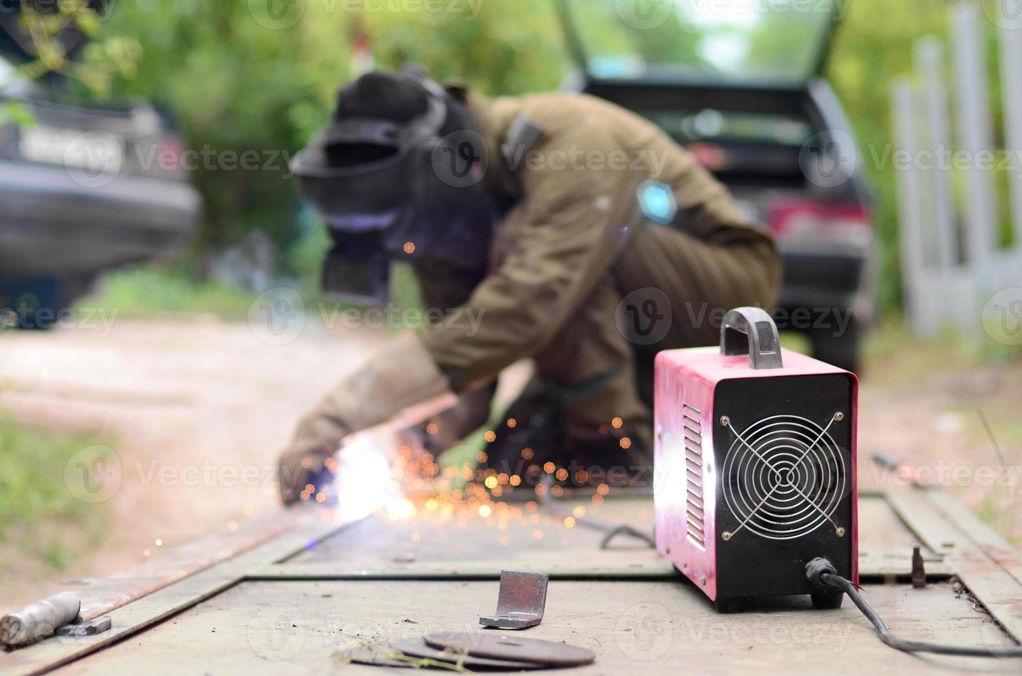 Old man welder in brown uniform, welding mask and welders leathers, weld metal door with arc welding machine outdoors photo