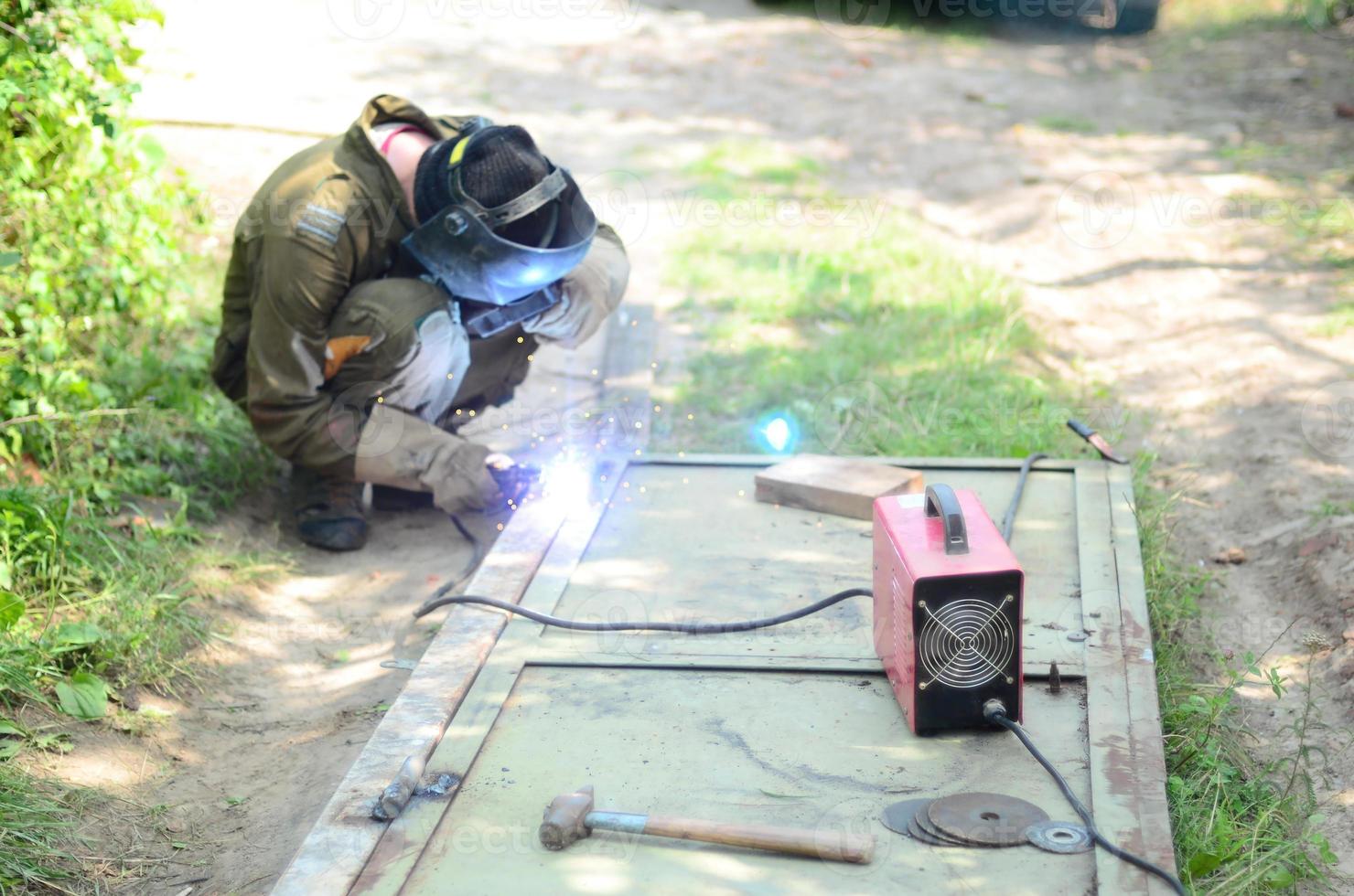 Old man welder in brown uniform, welding mask and welders leathers, weld metal door with arc welding machine outdoors photo