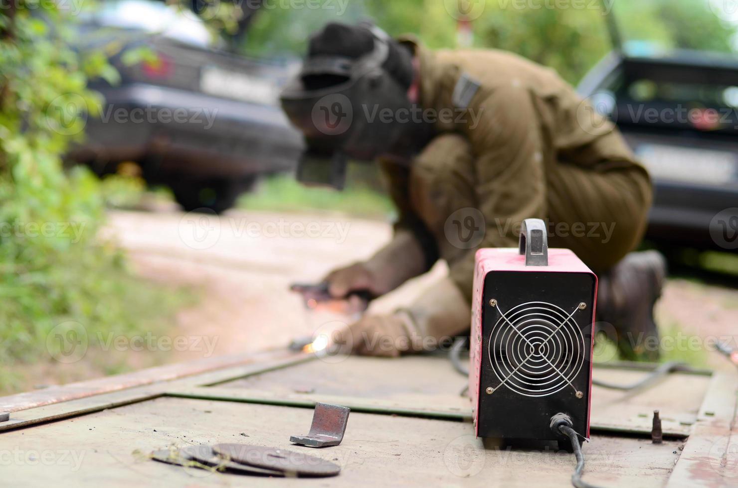 Old man welder in brown uniform, welding mask and welders leathers, weld metal door with arc welding machine outdoors photo