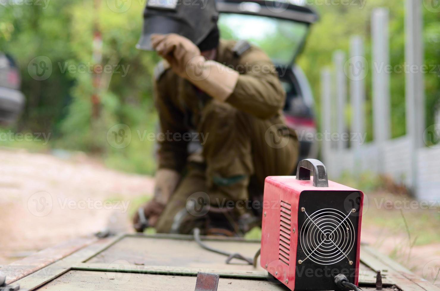 Old man welder in brown uniform, welding mask and welders leathers, weld metal door with arc welding machine outdoors photo