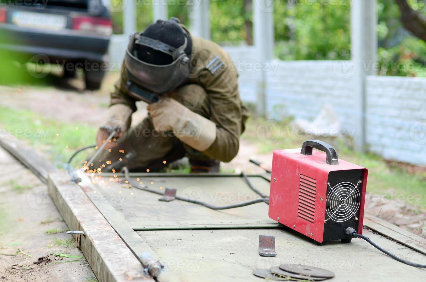 Old man welder in brown uniform, welding mask and welders leathers, weld metal door with arc welding machine outdoors photo