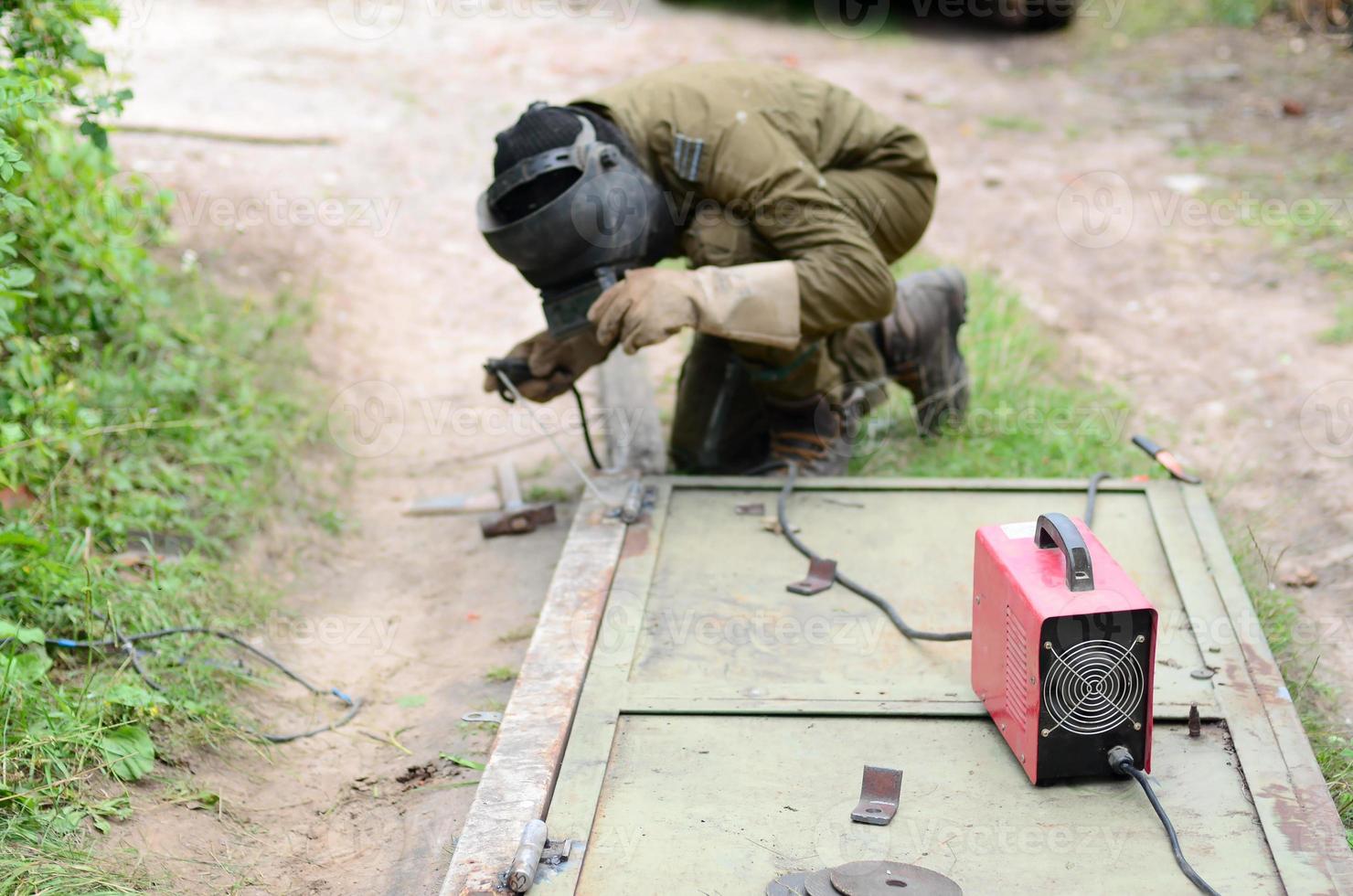 Old man welder in brown uniform, welding mask and welders leathers, weld metal door with arc welding machine outdoors photo