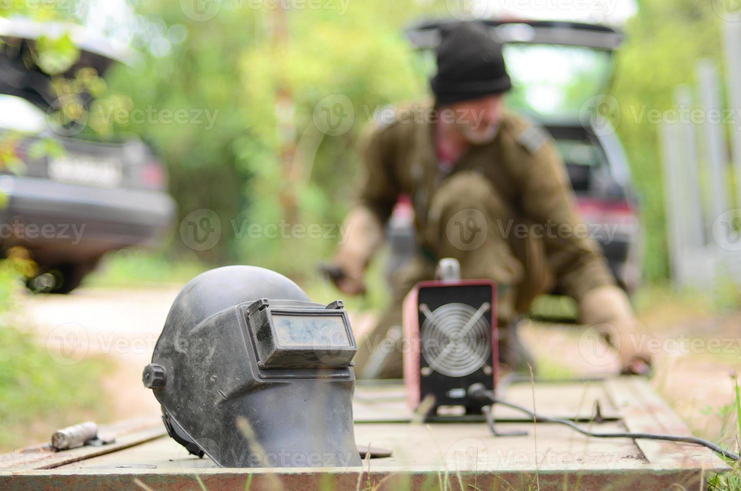 Old man welder in brown uniform prepares metal door surface for welding with arc welding machine outdoors photo