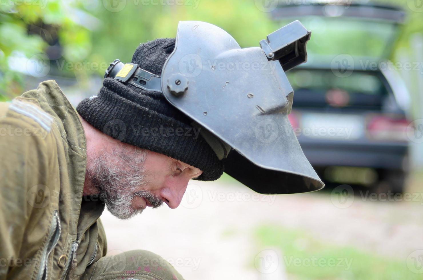 Old man welder in brown uniform prepares metal door surface for welding with arc welding machine outdoors photo