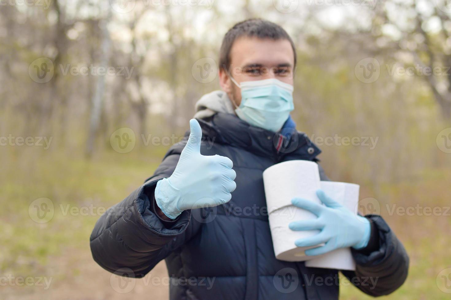 Covidiot concept. Young man in protective mask holds many rolls of toilet paper and shows thumb up outdoors in spring wood photo