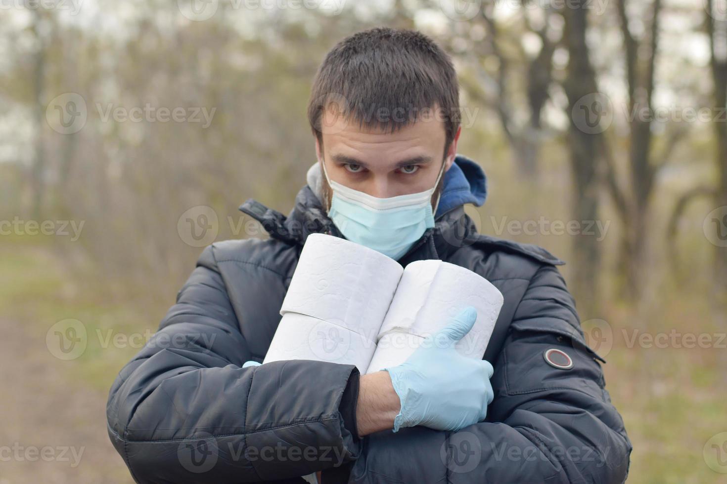 Covidiot concept. Young man in protective mask holds many rolls of toilet paper outdoors in spring wood photo