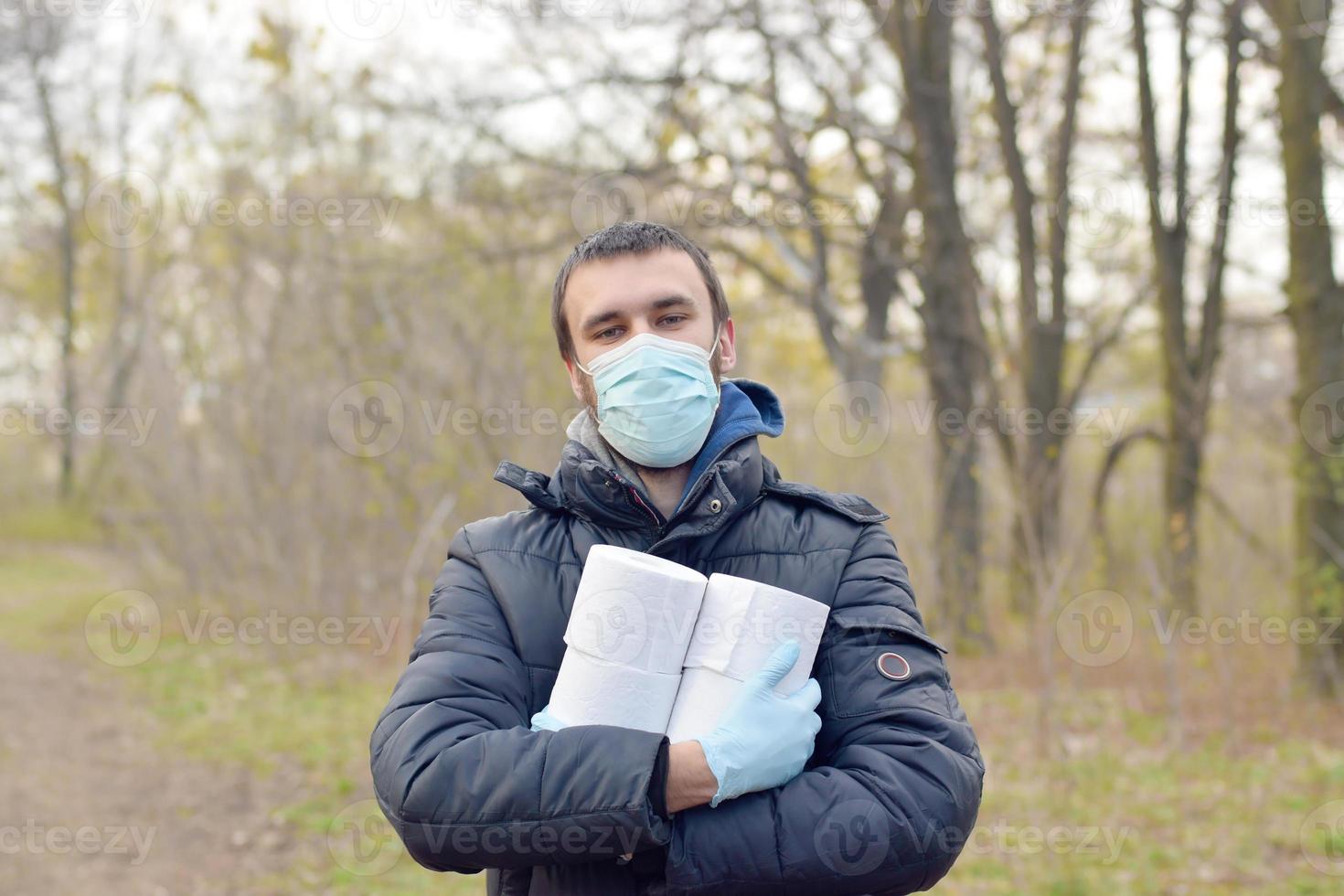 Covidiot concept. Young man in protective mask holds many rolls of toilet paper outdoors in spring wood photo