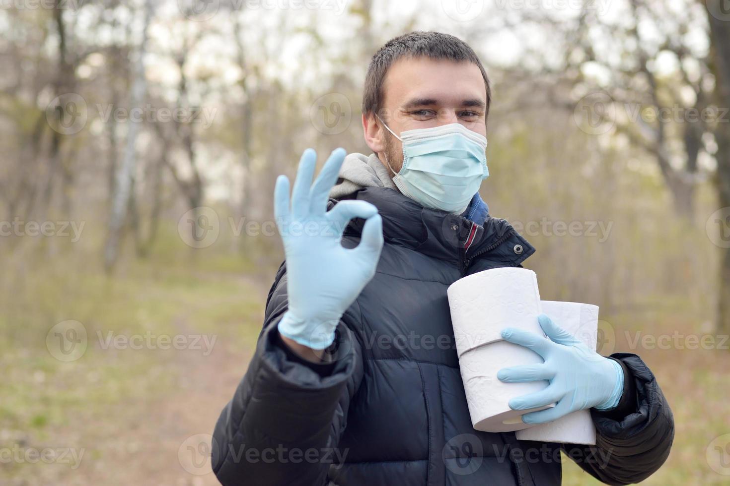 Covidiot concept. Young man in protective mask holds many rolls of toilet paper and shows okay gesture outdoors in spring wood photo