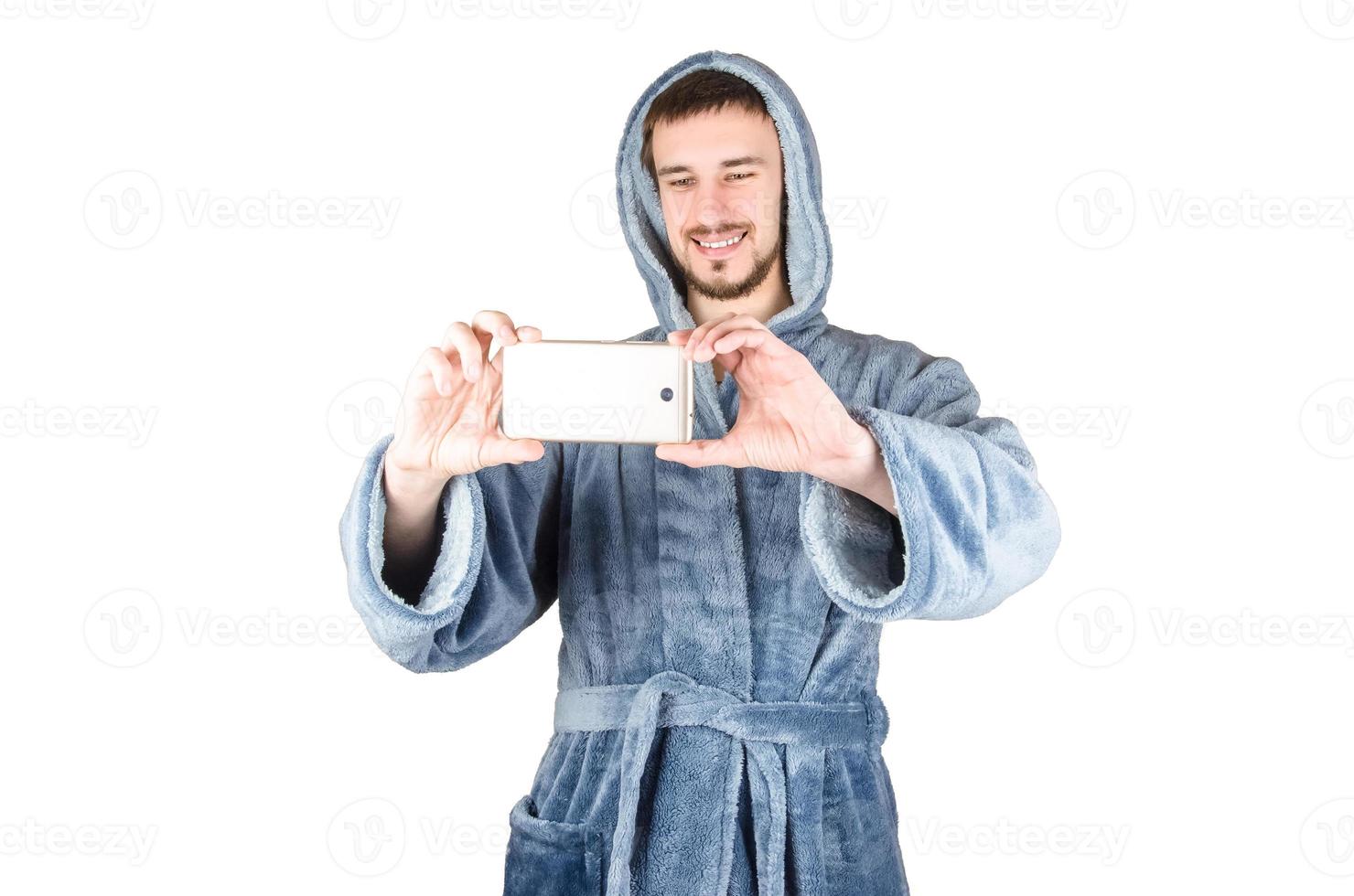 Portrait of young caucasian bearded man in blue bathrobe takes photo with smartphone isolated on white background