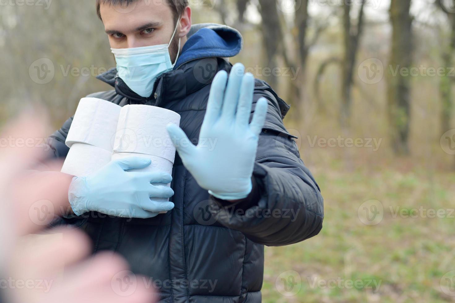 Covidiot concept. Young man in protective mask holds many rolls of toilet paper and shows stop gesture outdoors in spring wood photo