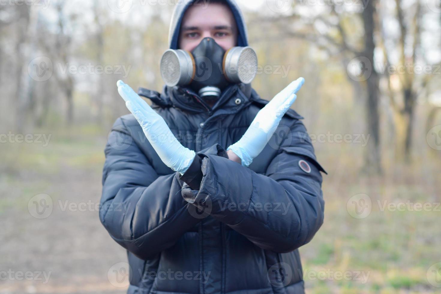 Portrait of young man in protective gas mask and rubber disposable gloves shows stop gesture outdoors in spring wood photo
