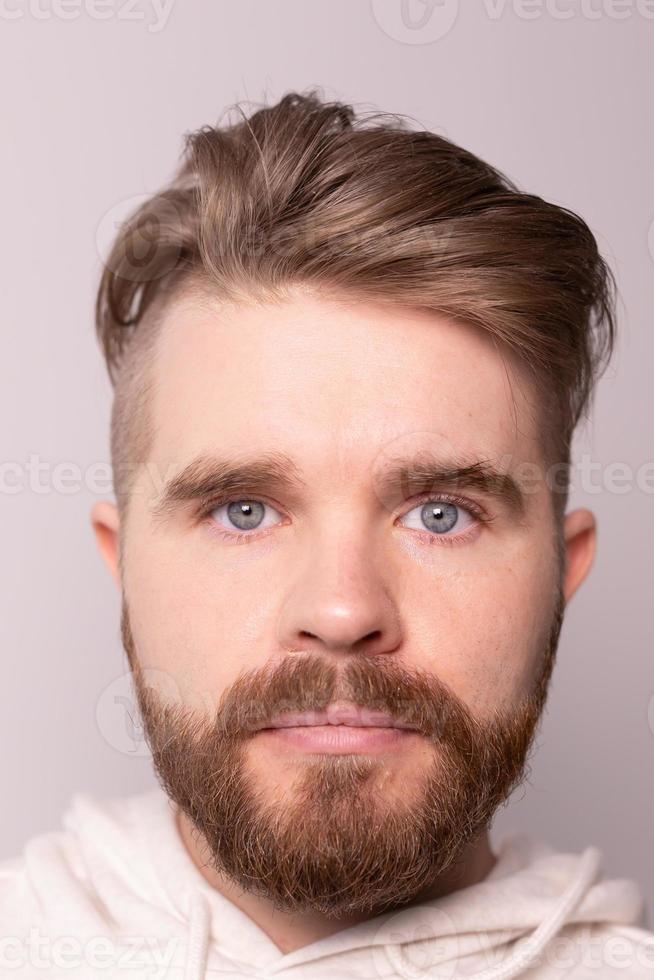 Close-up portrait of young male with beard and mustache and trendy hairdo. Wears casual hoodie, has serious expression in studio against white background photo