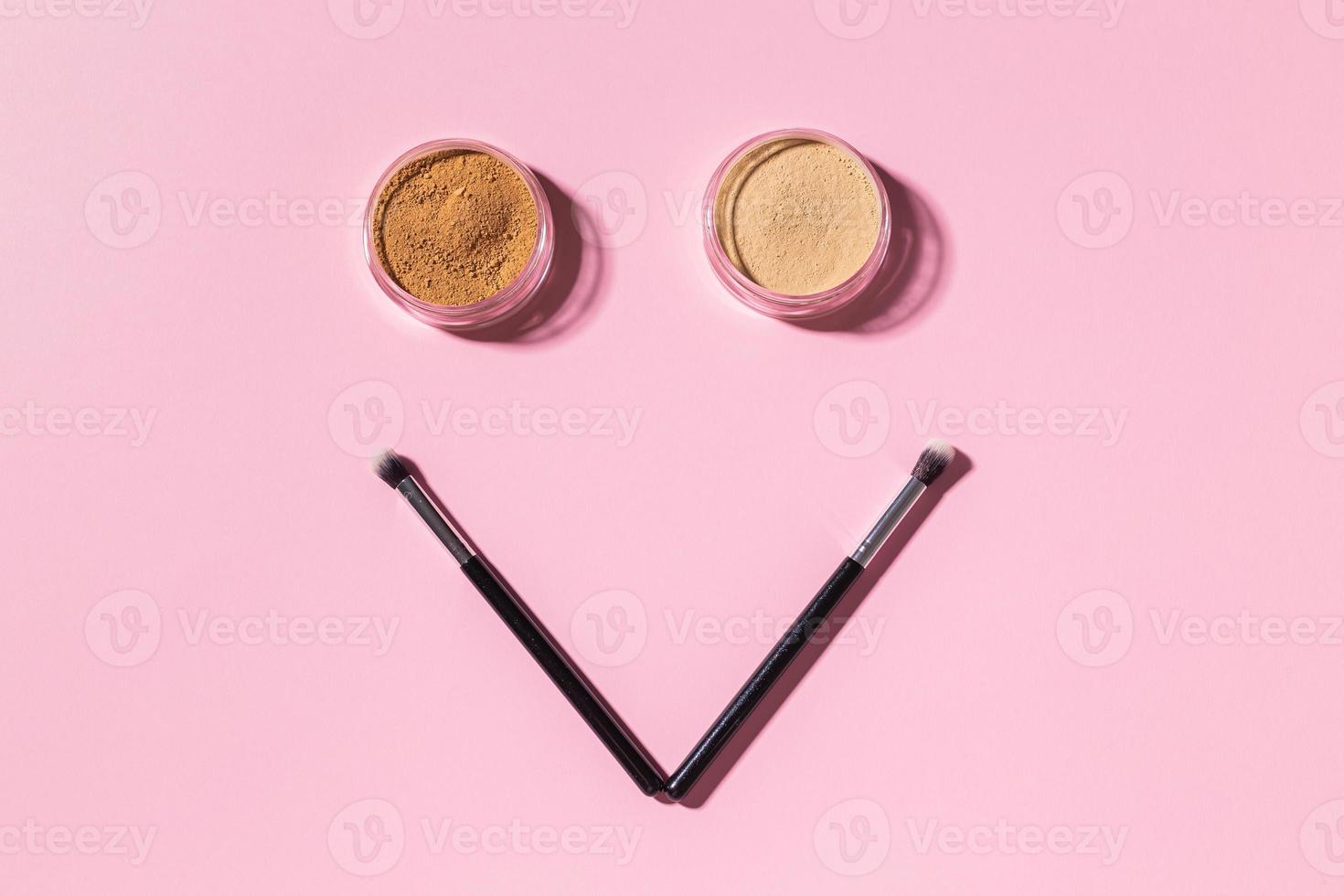 Funny face with make-up brushes and mineral powder on pink background with copy space, top view. Cosmetics humor and beauty concept. photo