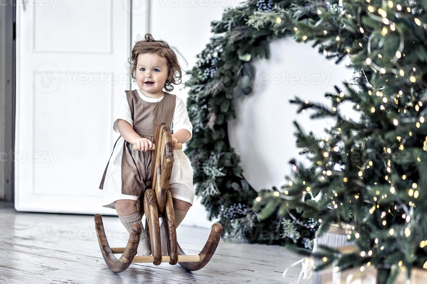 A little girl swings on a wooden horse in the room against the background of Christmas decorations. photo