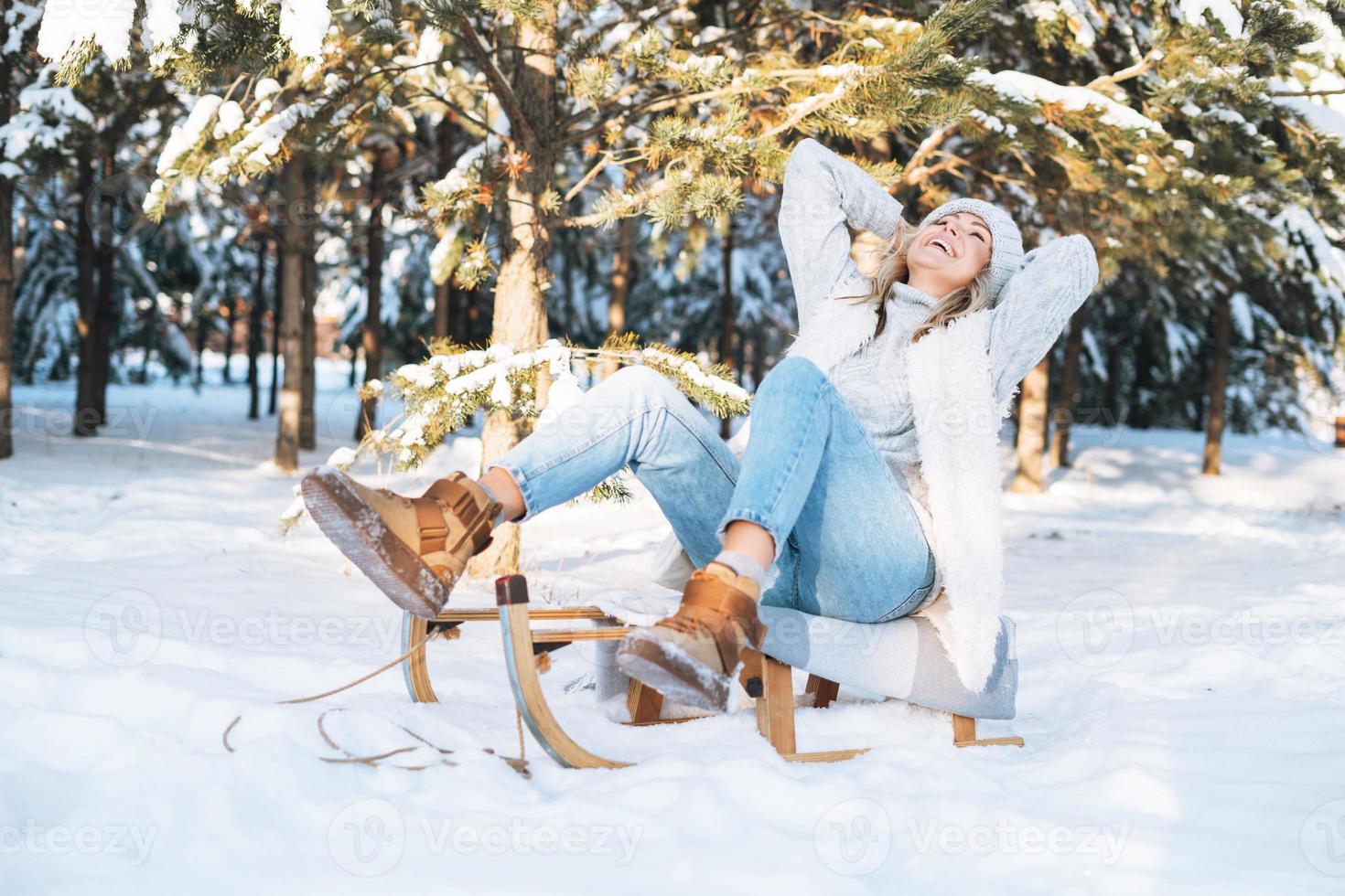 mujer feliz con cabello rubio en ropa de invierno divirtiéndose con trineo contra el fondo del bosque de invierno nevado foto