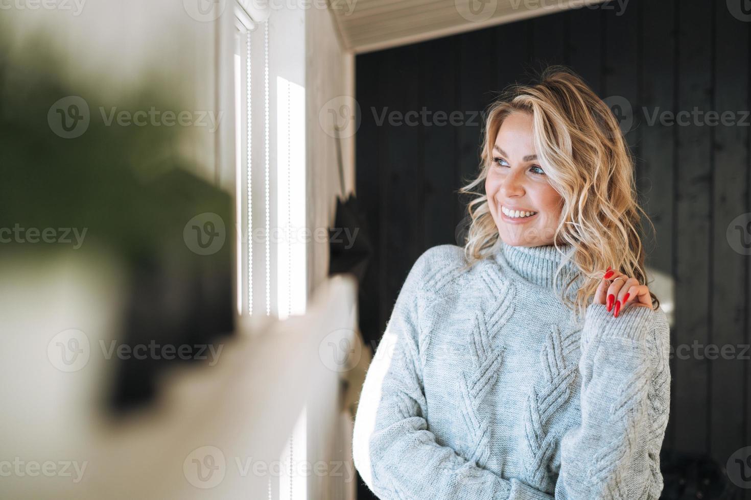 retrato de una mujer sonriente pensante de cuarenta años con el pelo rizado rubio en un suéter de punto gris cerca de la ventana de la casa foto