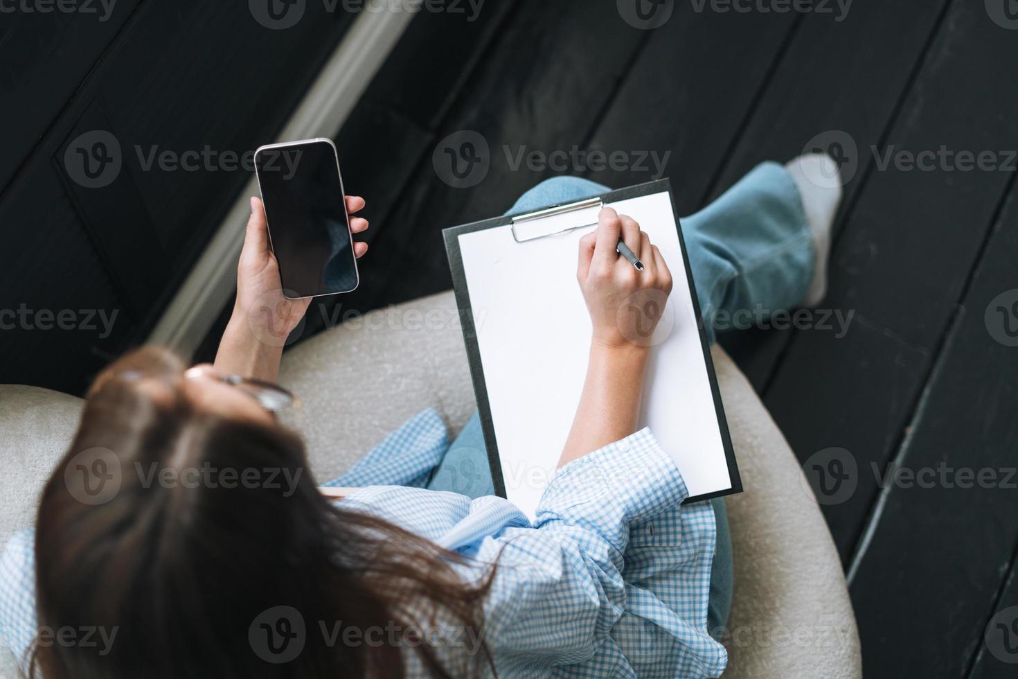 Crop photo of young woman in bue shirt with notes in hands using mobile phone on chair in room