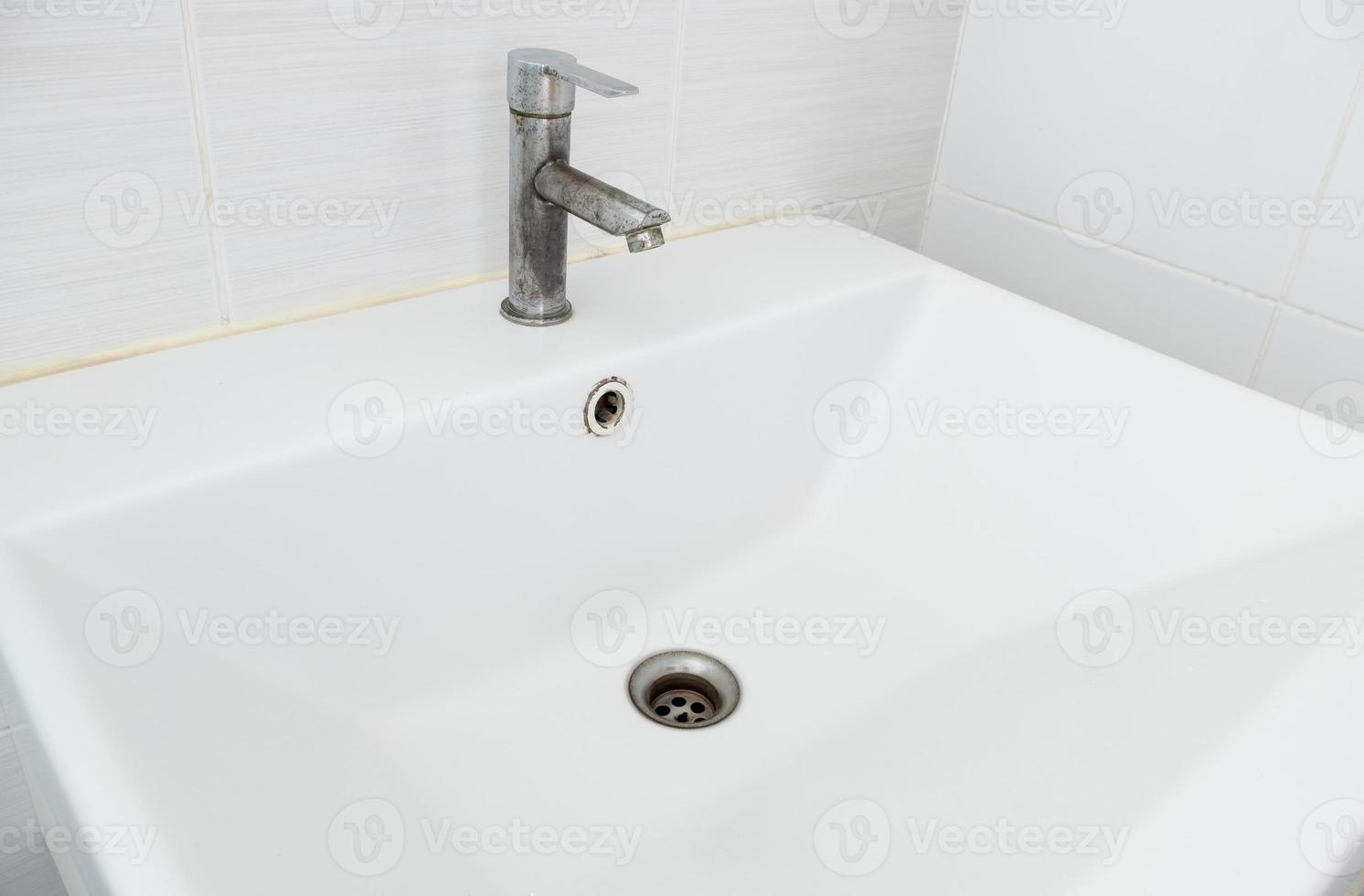 The ceramic sink basin with the stained faucet near the white tile wall. photo