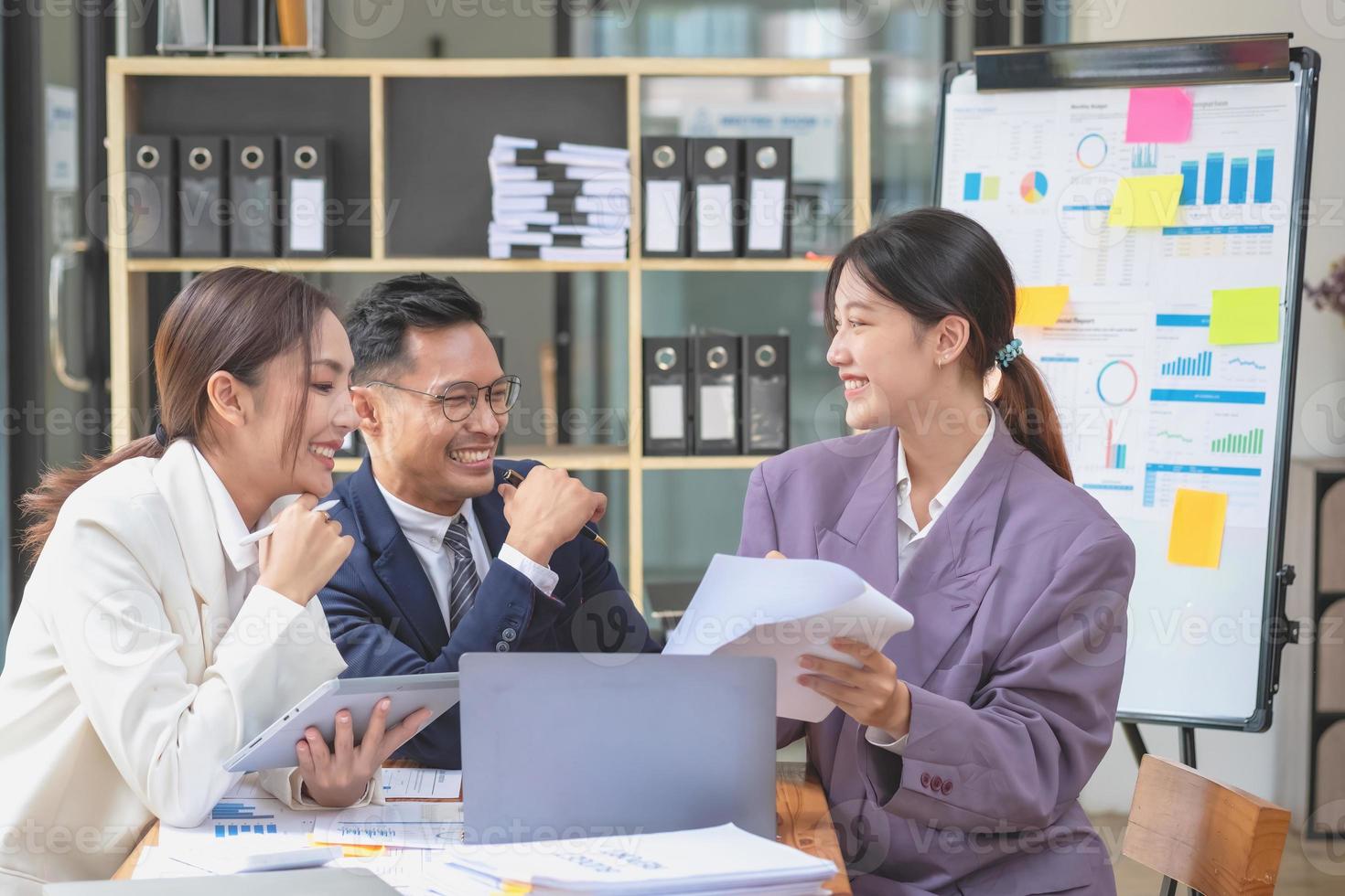 Team business three smiling and happy Asian men and women team using tablets and laptops brainstorm and plot statistics on a corporate chart board inside the company. Colleagues concept. photo