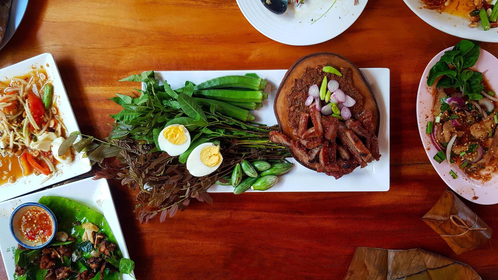 Flat lay of Thai food with fried pork,  chili paste with fermented fish and fresh vegetable. papaya salad, boiled egg, spicy grilled pork neck salad and sticky rice in banana leaf on wooden table. photo