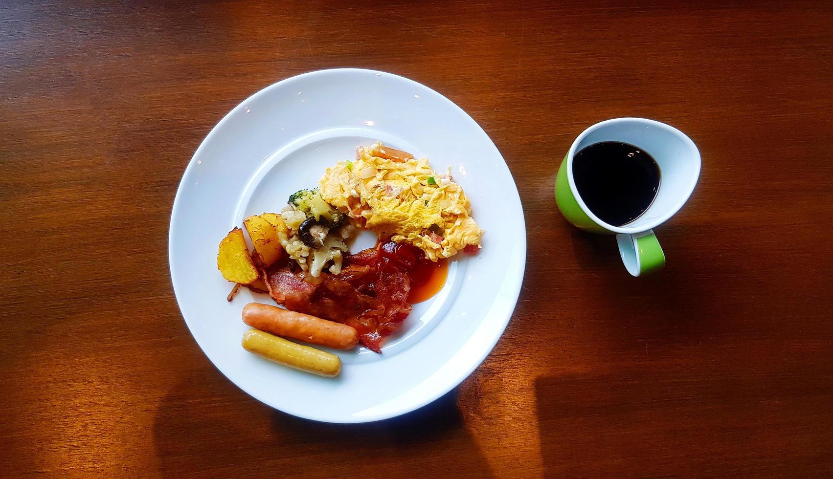 Top view of breakfast with omelet, bacon, many sausage, fried vegetable and tomato or chili sauce with green cup of black coffee. Delicious meal in American style with hot drinking on wooden table. photo
