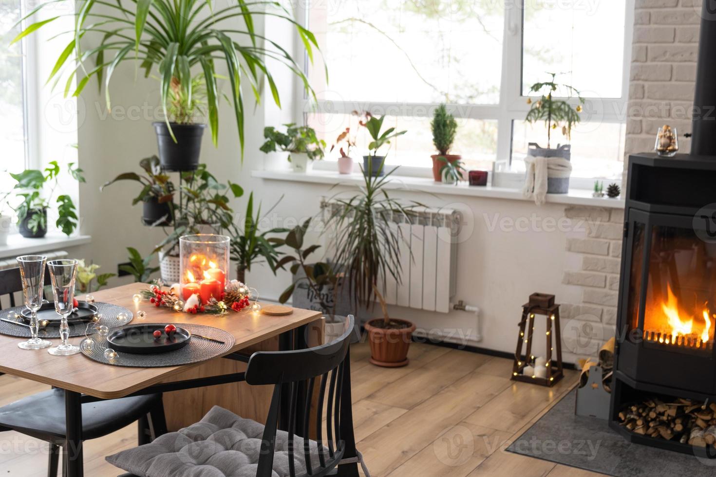 el interior festivo de la casa está decorado para navidad y año nuevo en estilo loft con estufa negra, chimenea, árbol de navidad. cálida sala de estudio con mesa puesta, madera quemada, acogedora y calefacción del hogar foto