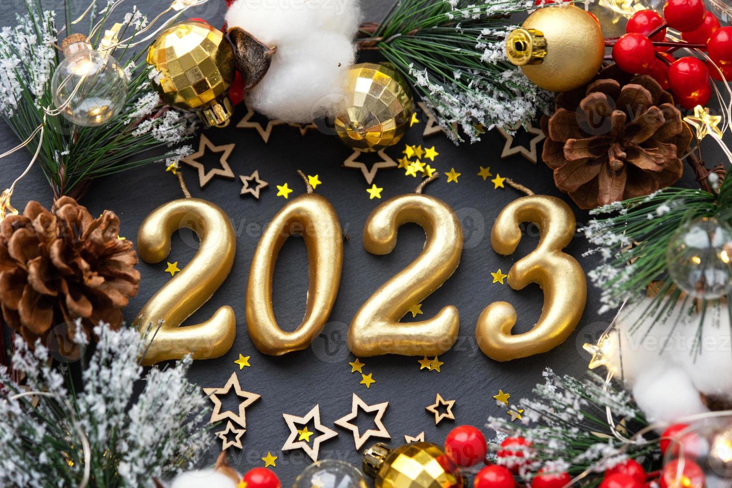 The golden figures 2023 made of candles on a black stone slate background are decorated with a festive decor of stars, sequins, fir branches, balls and garlands. Greeting card, happy New Year. photo