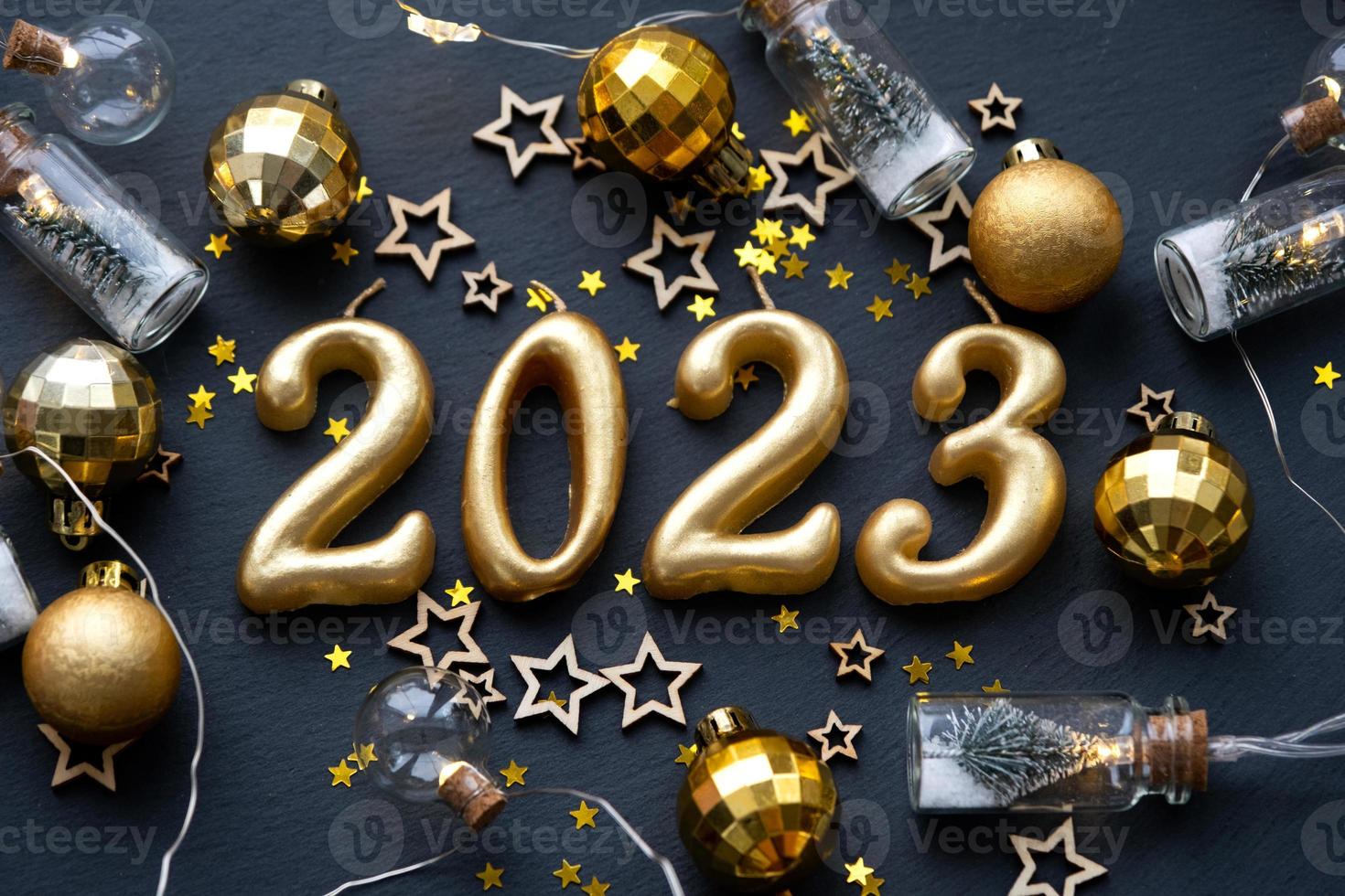 The golden figures 2023 made of candles on a black stone slate background are decorated with a festive decor of stars, sequins, fir branches, balls and garlands. Greeting card, happy New Year. photo