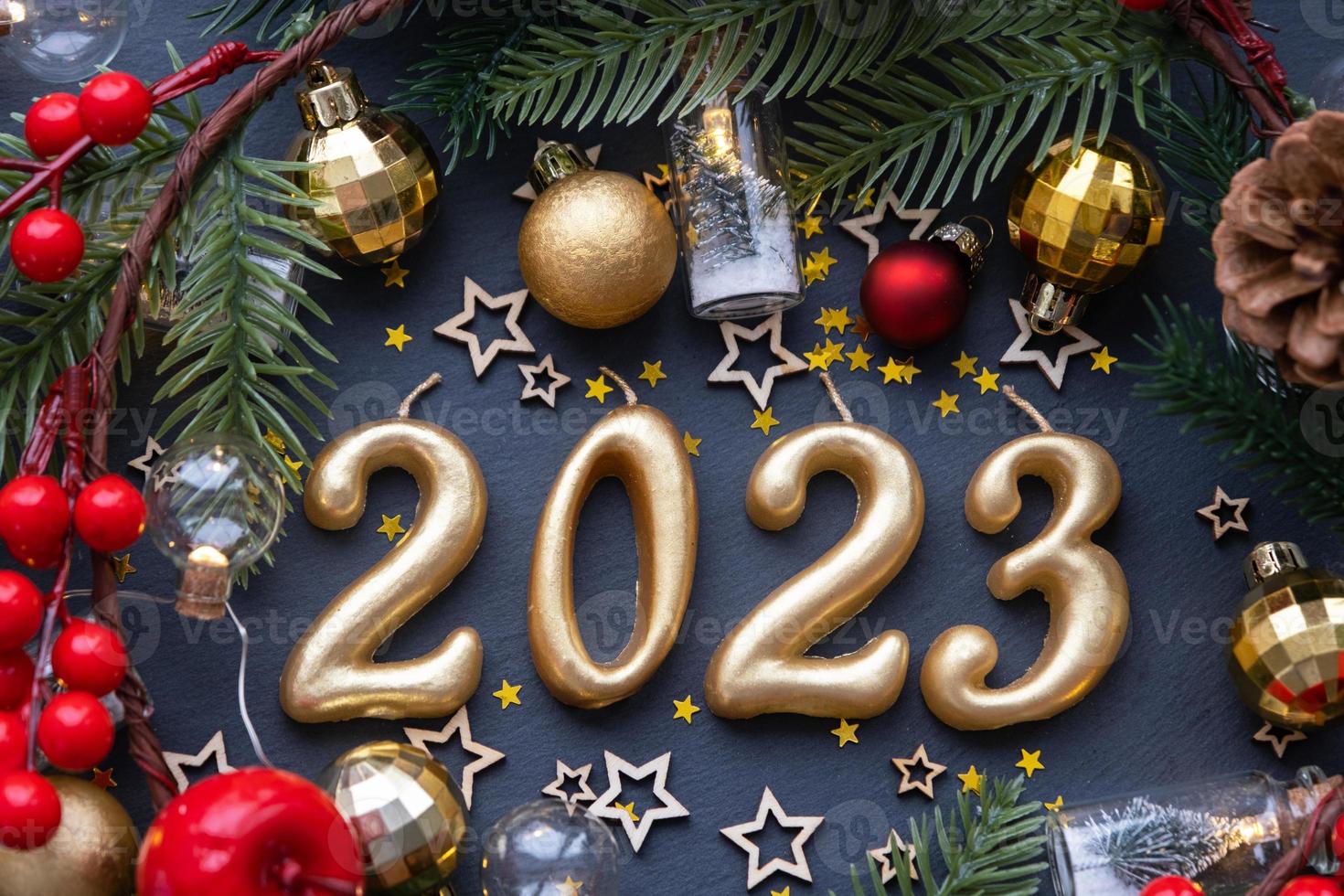 The golden figures 2023 made of candles on a black stone slate background are decorated with a festive decor of stars, sequins, fir branches, balls and garlands. Greeting card, happy New Year. photo