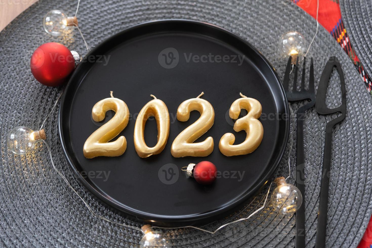 figuras doradas 2023 de velas en un plato negro en una mesa festiva con una porción de año nuevo. interior estilo loft, fiesta, fiesta. servilleta de mimbre, tenedor, cuchillo foto