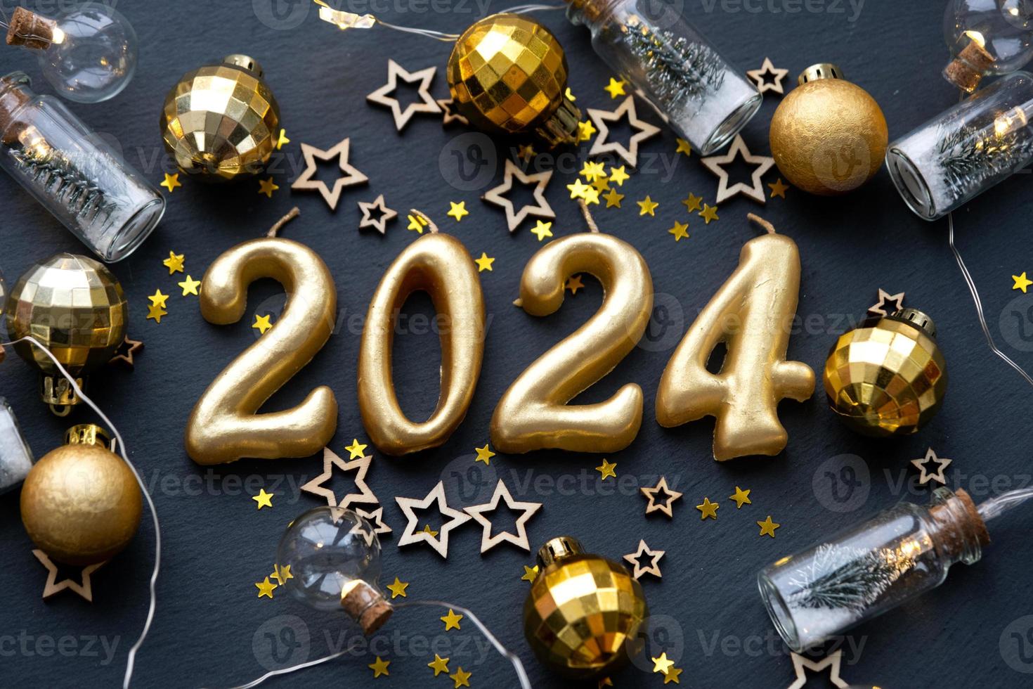 The golden figures 2024 made of candles on a black stone slate background are decorated with a festive decor of stars, sequins, fir branches, balls and garlands. Greeting card, happy New Year. photo