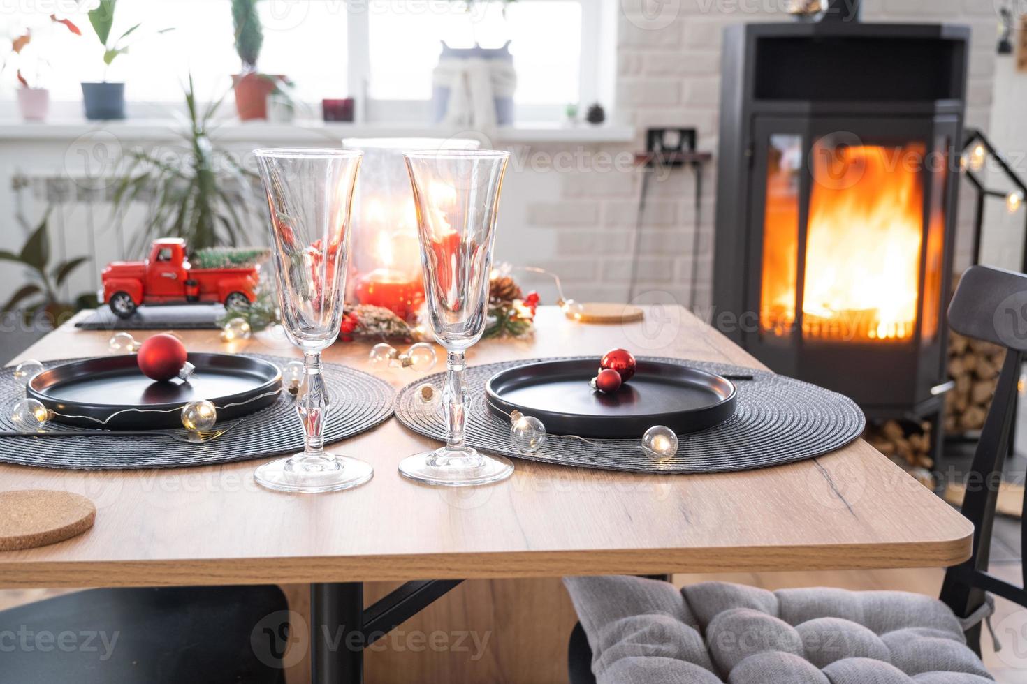 Festive interior of house is decorated for Christmas and New Year in loft style with black stove, fireplace, Christmas tree. Warm studio room with set table, burning wood, cozy and heating of home photo
