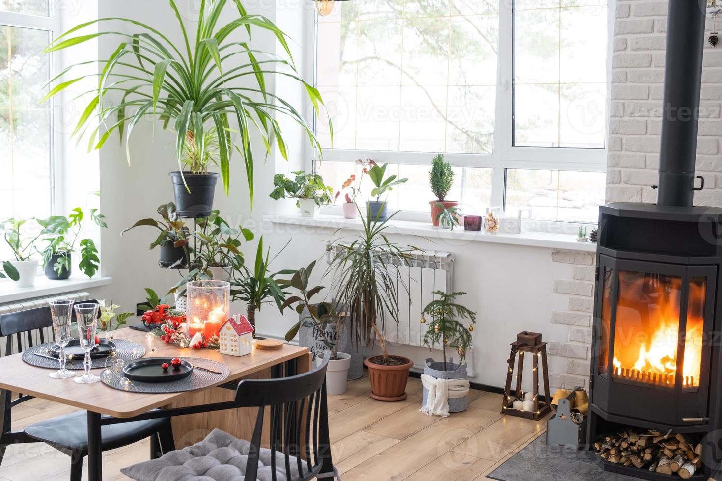 el interior festivo de la casa está decorado para navidad y año nuevo en estilo loft con estufa negra, chimenea, árbol de navidad. cálida sala de estudio con mesa puesta, madera quemada, acogedora y calefacción del hogar foto