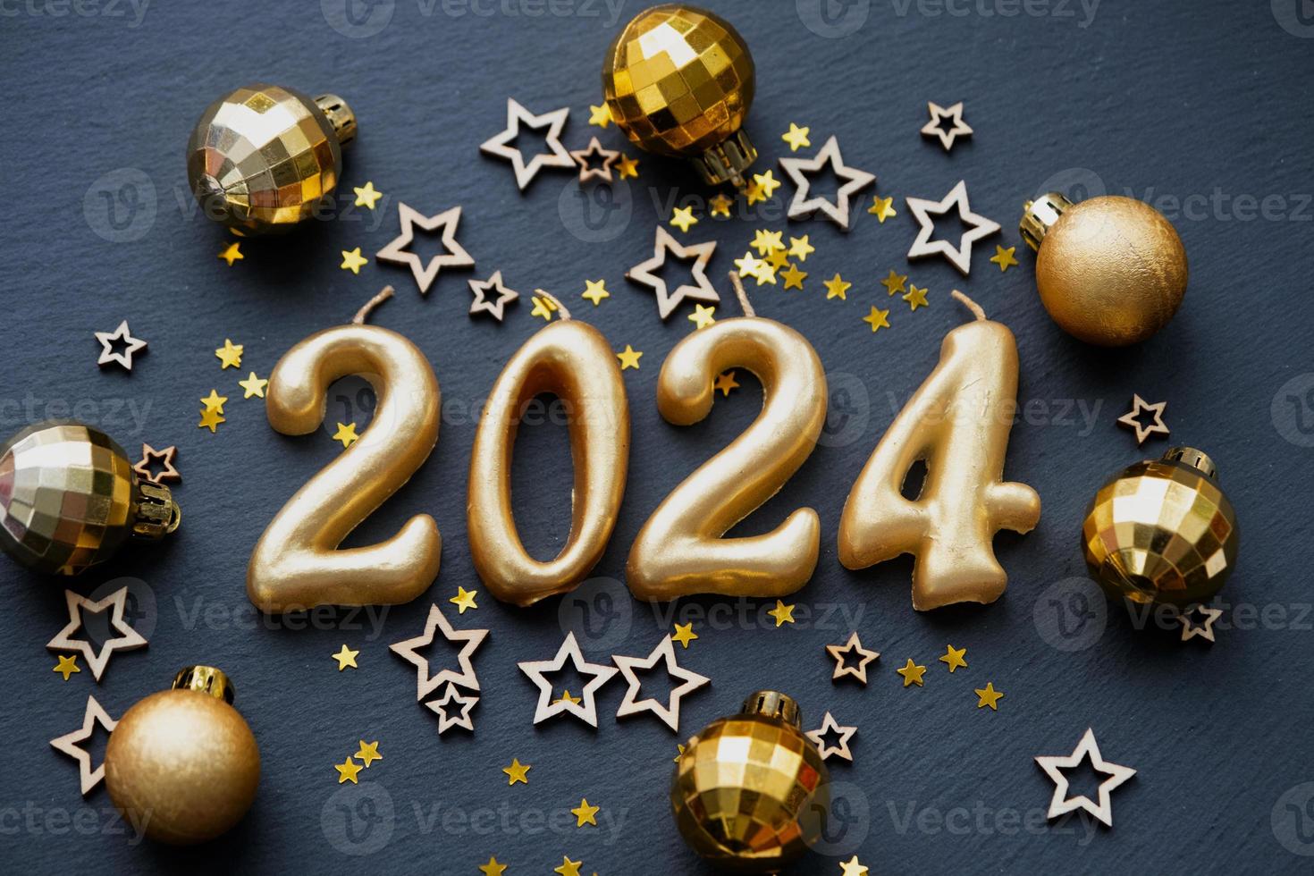 The golden figures 2024 made of candles on a black stone slate background are decorated with a festive decor of stars, sequins, fir branches, balls and garlands. Greeting card, happy New Year. photo