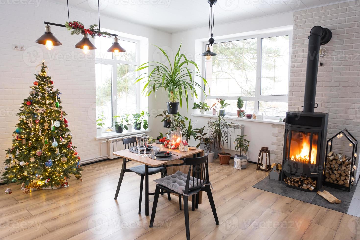 el interior festivo de la casa está decorado para navidad y año nuevo en estilo loft con estufa negra, chimenea, árbol de navidad. cálida sala de estudio con mesa puesta, madera quemada, acogedora y calefacción del hogar foto