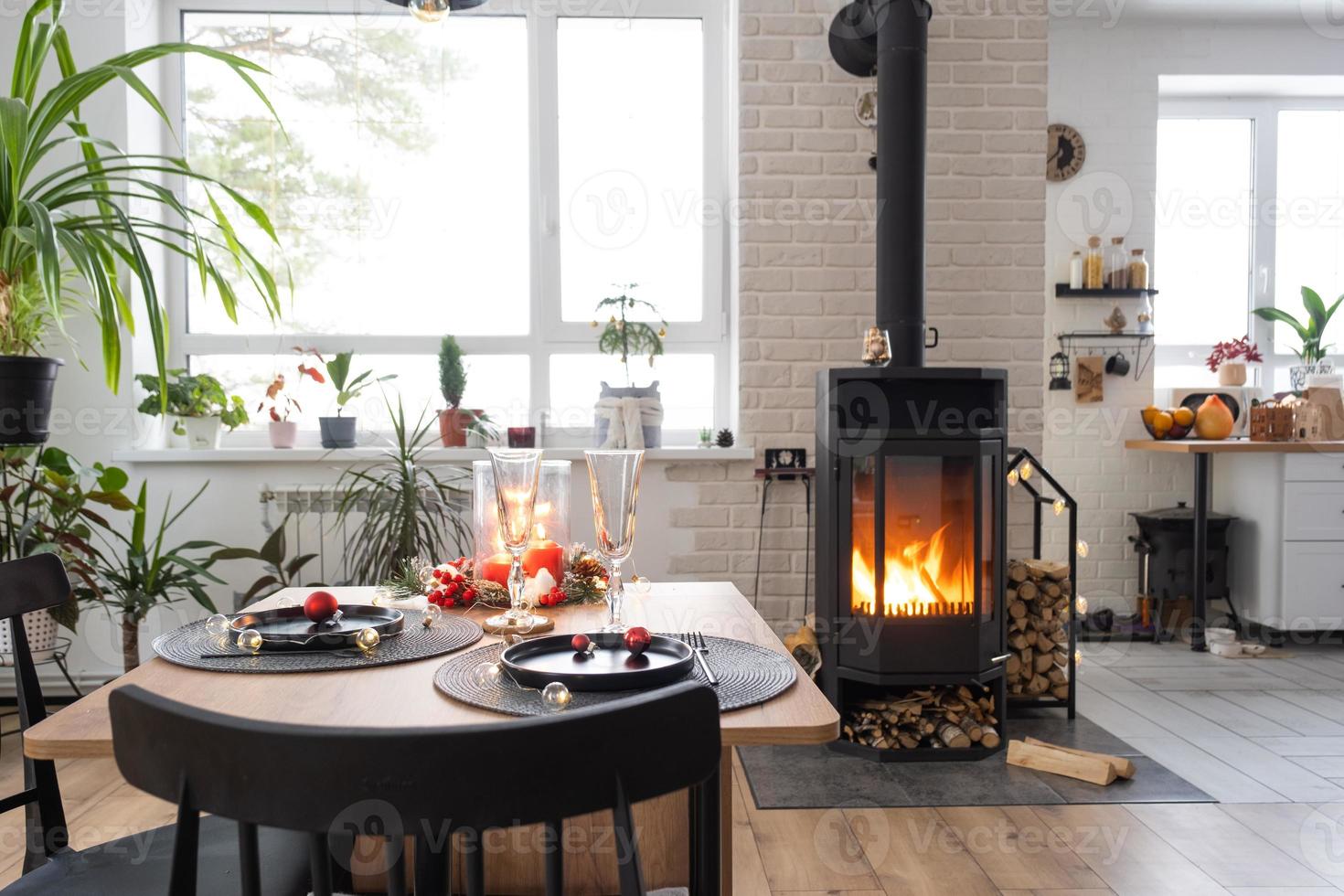 Festive interior of house is decorated for Christmas and New Year in loft style with black stove, fireplace, Christmas tree. Warm studio room with set table, burning wood, cozy and heating of home photo